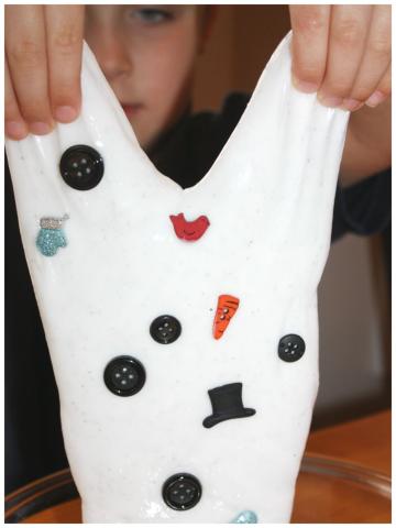 Child stretching white slime with pieces of a snowman mixed in such as a tiny carrot nose, buttons and a miniature top hat