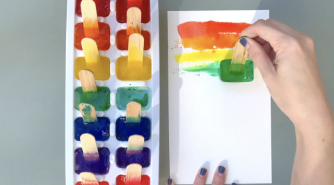 ice tray filled with colorful paint ice cubes, hands using a green ice paint pop to make lines on paper