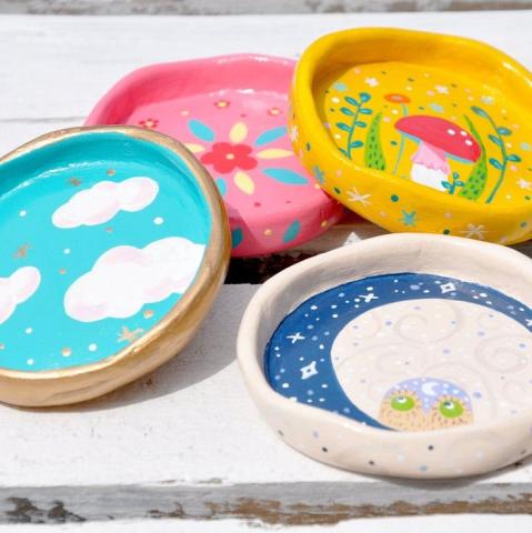 four assorted colorful round trinket dishes. Two have a red capped mushroom on them. One of these is pink, one is yellow. Another dish is blue with white clouds on it. The last one has a dark blue crescent moon.