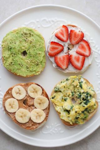 four half bagels on the plate: one with avocado spread, one with sliced strawberries and cream cheese, one with peanut butter and sliced banana and one with a scrambled egg