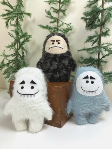 three small yeti plushes: one white, one brown, one steel blue. They have prominent eyebrows and the brown one is frowning 