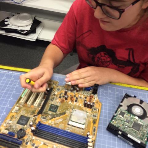 photograph of a young person wearing glasses and holding a small screwdriver as they tinker with an unidentified piece of technology